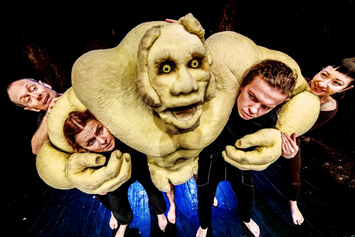 Bajkowa postać ze spektaklu, obejmująca ramionami dwóch aktorów, za nią kolejne dwie osoby.