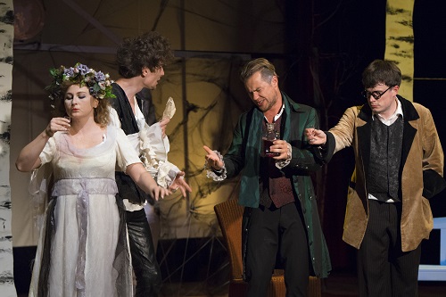 Trzech mężczyzn i jedna kobieta na scenie. Kobieta ubrana w białą suknię z wiankiem na głowie trzyma w dłoni kieliszek, mężczyźni żywo o czymś dyskutują.