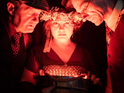 Oświetlone twarze dwóch mężczyzn i kobiety w wianku na głowie. Twarze trojga ludzi są blisko siebie, patrzą na coś w dole.