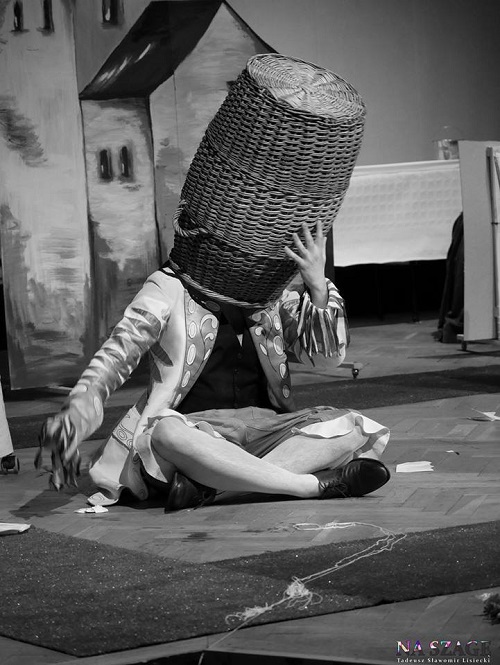 Czarno-białe zdjęcie osoby siedzącej na podłodze ze skrzyżowanymi nogami z dużym koszum na głowie; kosz całkowicie zakrywa twarz.