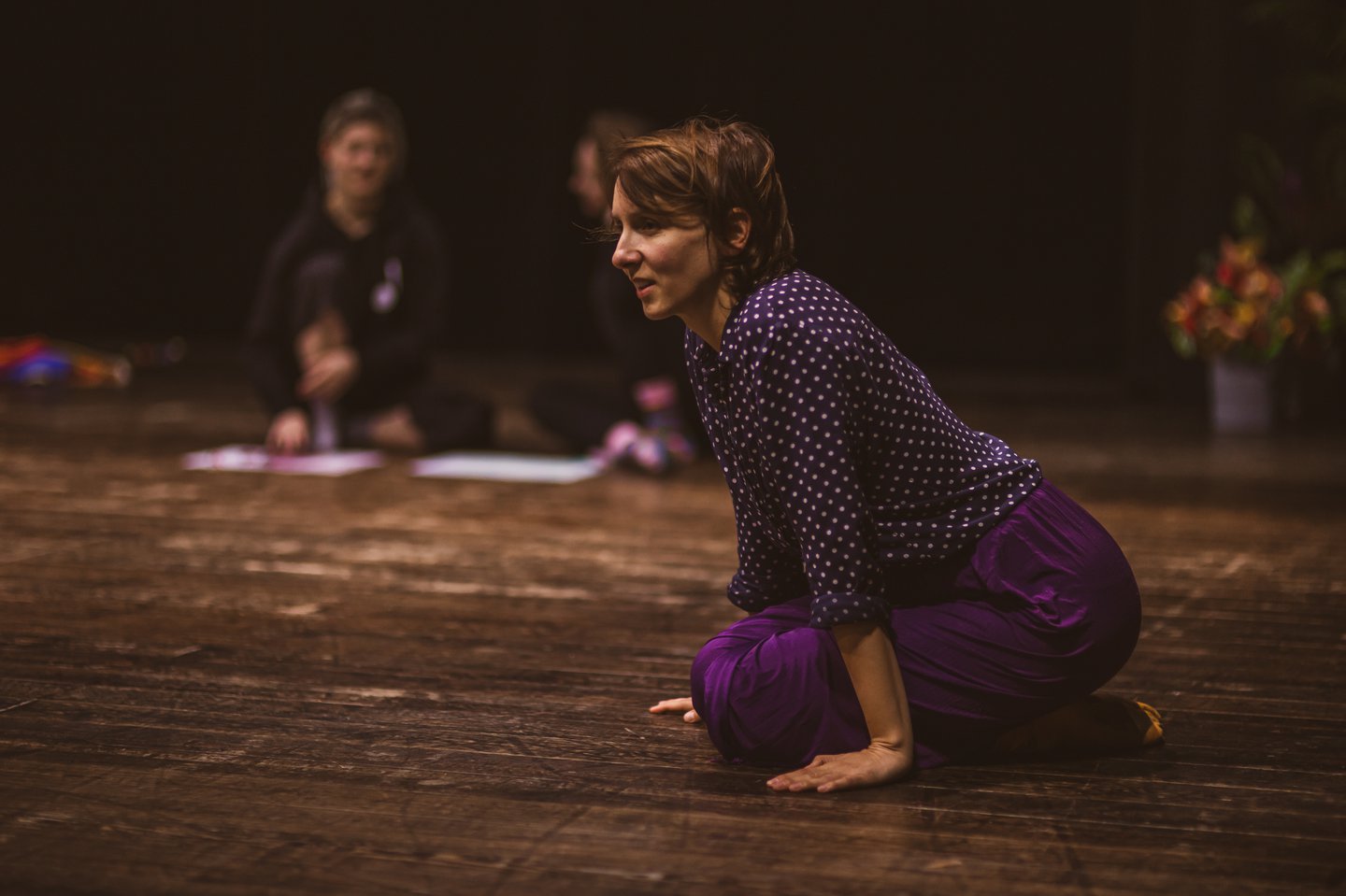 Na podłodze klęcząca kobieta, podpierająca się rękami. W tle dwie osoby, siedzące na podłodze z podkurczonymi nogami.