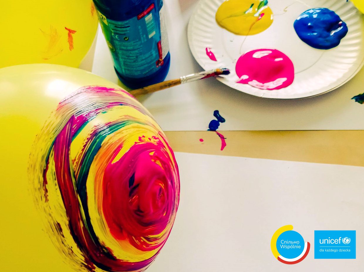 Kolorowe zdjęcie żółtego balona częściowo pomalowanego w kolorowe okręgi. Na drugim planie papierowy talerz z trzema kolorowymi plamami, o talerz oparty jest pędzelek.