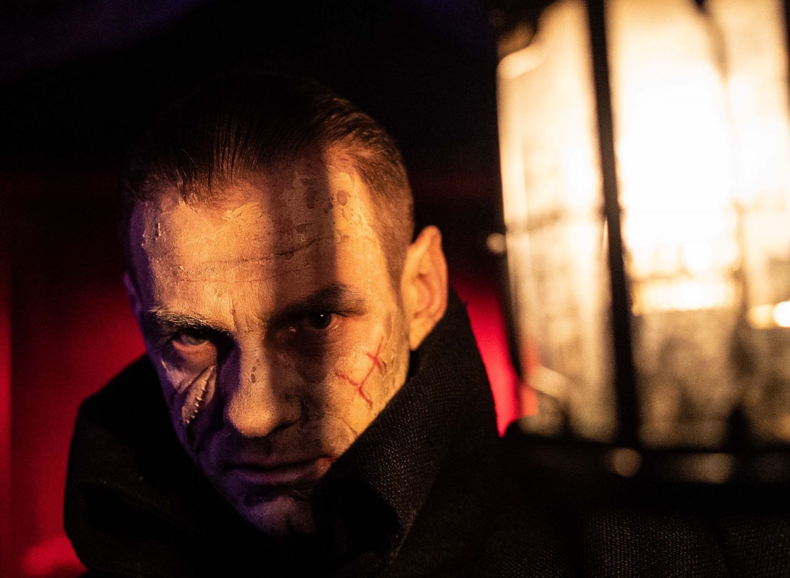 Twarz mężczyzny w półmroku, na którą częściowo pada światło. Na twarzy widoczne blizny.