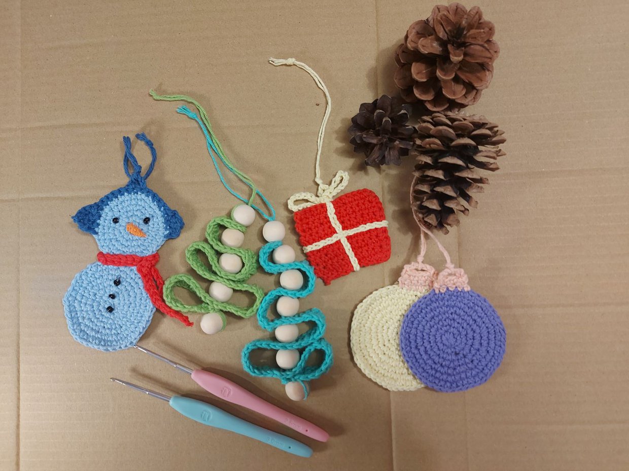 Na zdjęciu kilka prac z kolorowej włóczki wykonanych szydełkiem, takich jak niebieski bałwanek, bombki, choinki. Na zdjęciu też trzy szyszki i dwa szydełka.