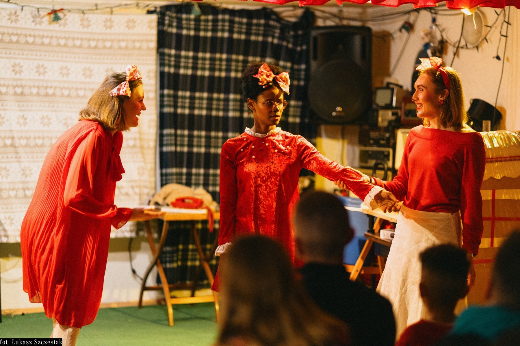 Trzy kobiety na scenie, są podobnie ubrane, każda z nich ma kokardę we włosach. W tle przedmioty codziennego użytku, w tym stacjonarny telefon na stoliku.