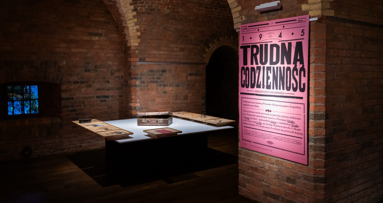 Wnętrze sali wystawowej w ciemnym piwnicznym pomieszczeniu. Na ścianie oświetlony plakat wystawy, w tle stół, na którym leży kilka przedmiotów, m.in. mały kuferek.