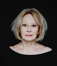 Zdjęcie kobiety w jasnych włosach, ubranej na czarno, na czarnym tle. Kobieta patrzy prosto w obiektyw.