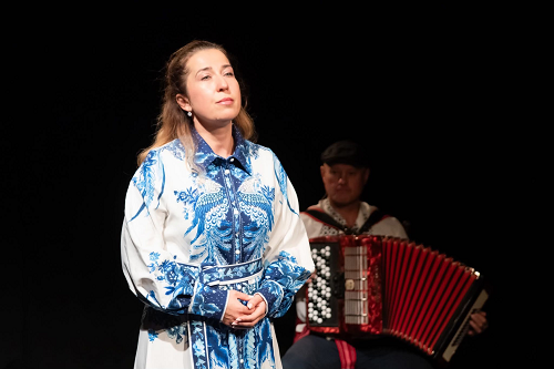 Zdjęcie kobiety w biało-niebieskiej sukience, która patrzy w dal. W tle mężczyzna grający na akordeonie.