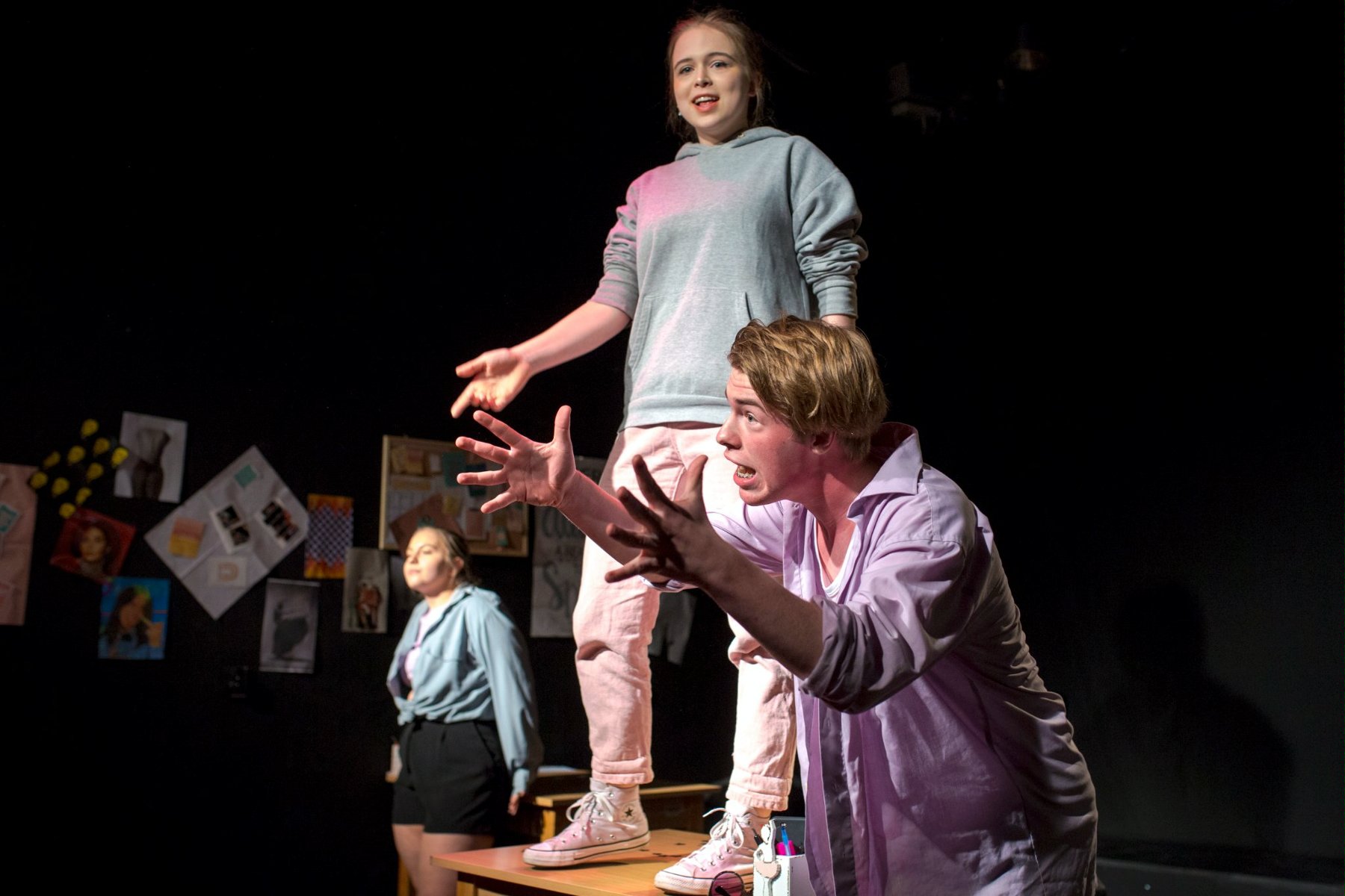 na zdjęciu troje młodych aktorów, na pierwszym planie młody mężczyzna pochylony do przodu gestykuluje rękami, za nim uśmiechnięta dziewczyna stoi na stole, w tle kolejna dziewczyna stoi oparta o stół