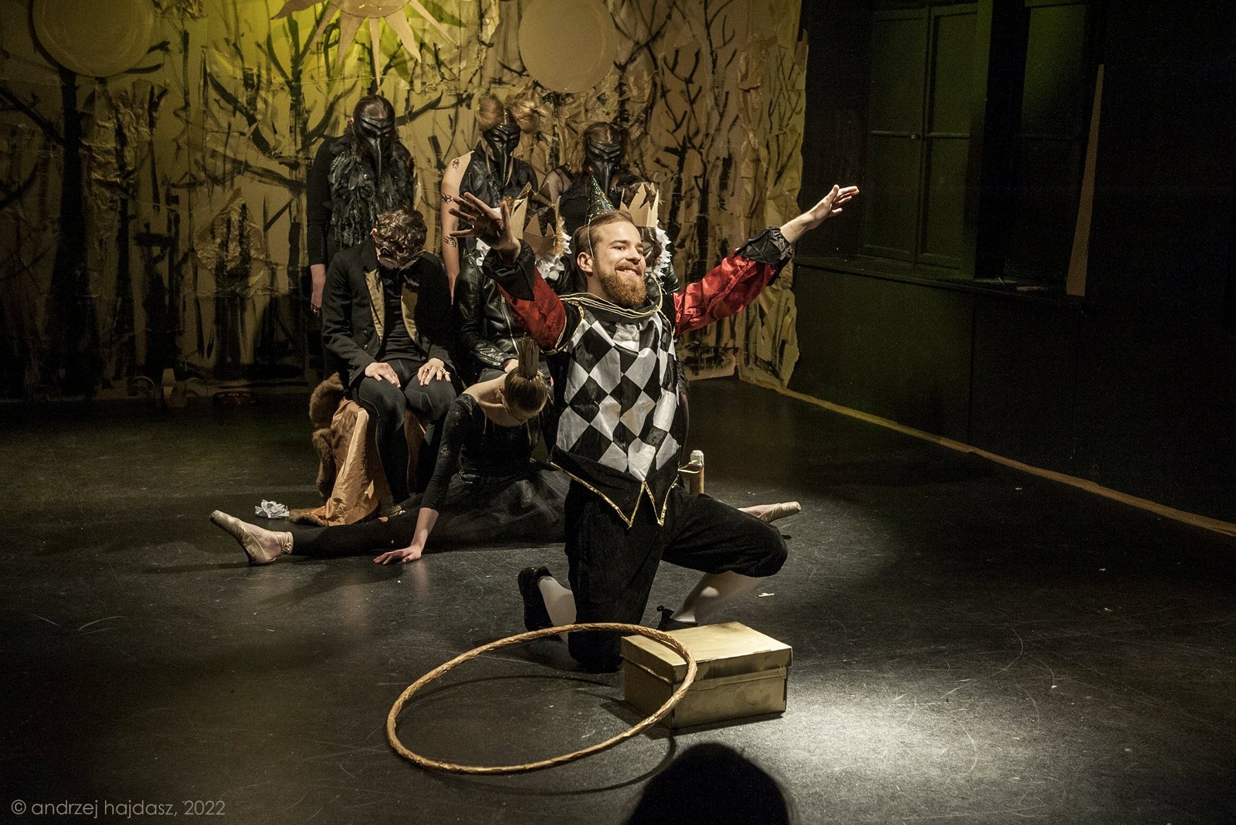 na pierwszym planie mężczyna klęczący na jedno kolano, przed nim stoi pudełko i leży hulahop. Mężczyzna ma kolorowy strój: czarne spodnie do kolan, białe kolanówki, czerwoną koszulę, kamizelkę w biało-czarną szachownicę. W tle kilka postaci ubranych na czarno w różnych pozach.