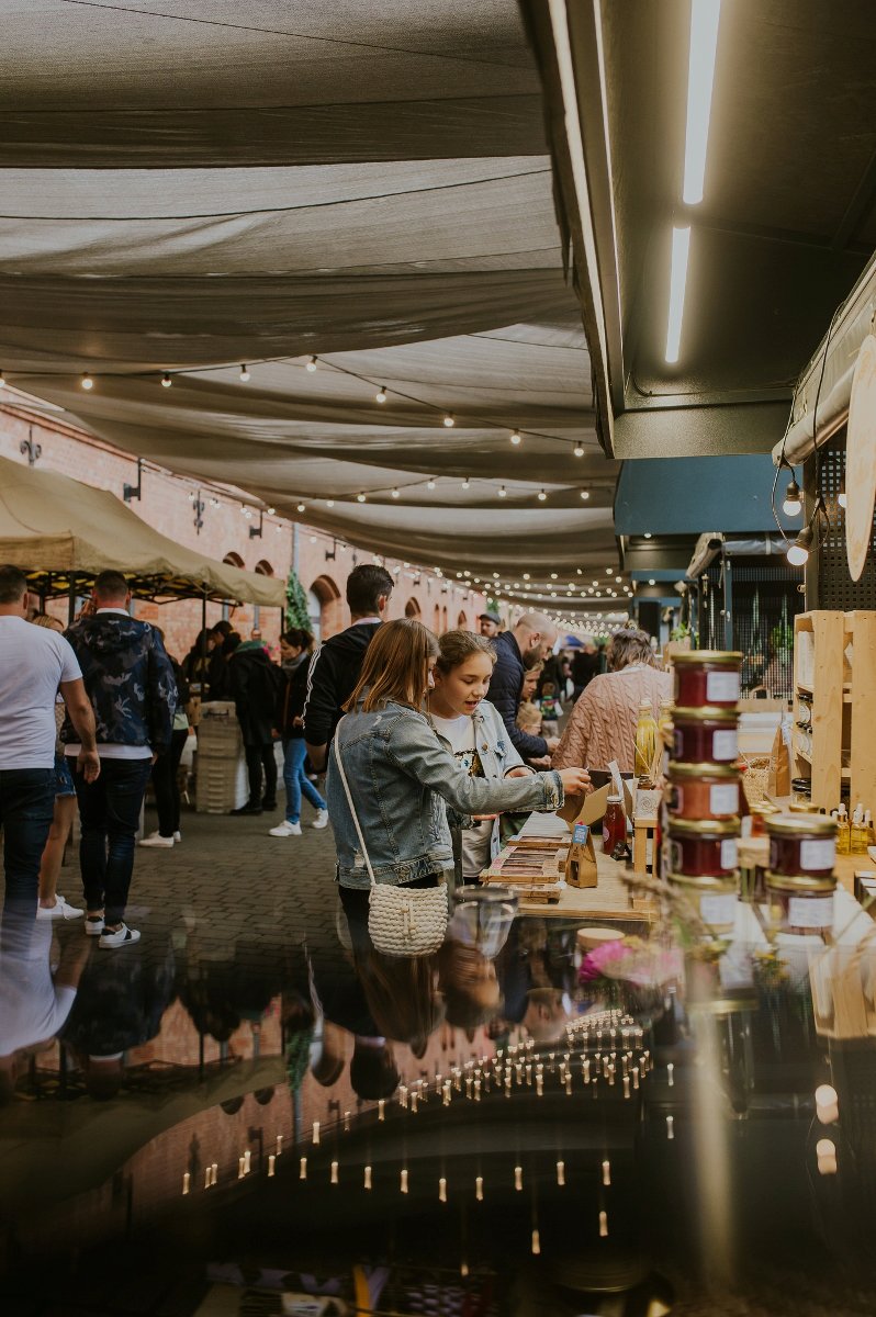 Ulica pod baldachimami pełna stoisk, a wśród nich chodzą ludzie i oglądają lub kupują wyroby rzemieślnicze.