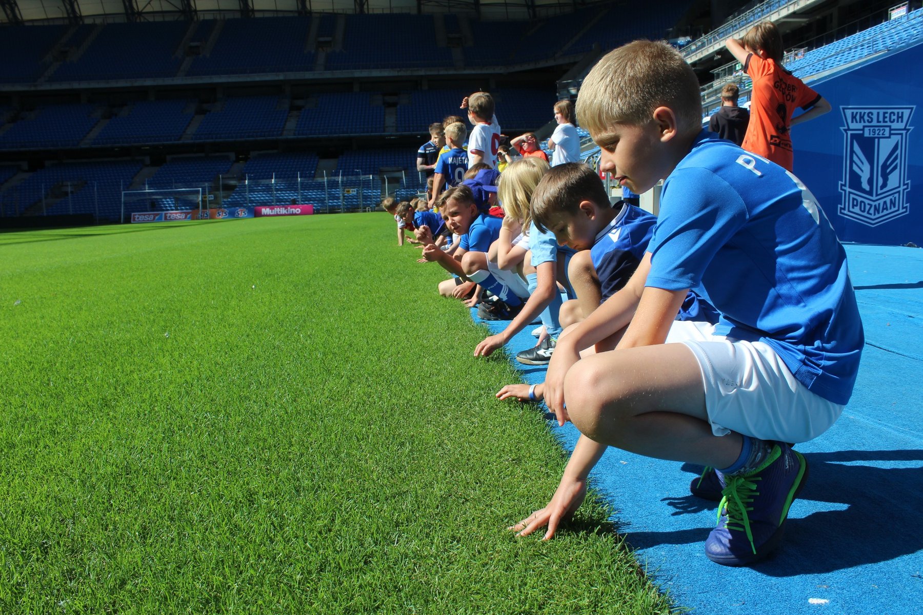 Zdjęcie przedstawiuczestników półkolonii, którzy w strojach piłkarskich siedzą przy boisku i dotykają trawy.