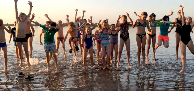 Na zdj eciu znajduje się grupa dzieci, która skaczę w jeziorze z podniesionymi ku górze rękoma.