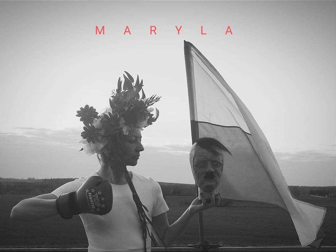 Czarno-białe zdjęcie kobiety z kwiecistym wiankiem na głowie, która w jednej ręce trzyma rzeźbę - głowę mężczyzny, a na drugiej ręce ma rękawicę bokserską w pozie do uderzenia. Za kobietą flaga, w tle otwarta przestrzeń.