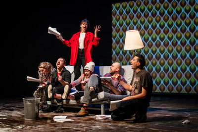 na kanapie na scenie siedzi kilka osób, niektórzy trzymają w rękach otwarte książki. Za nimi stoi kobieta z otwartą książką w ręce, mocno gestykuluje