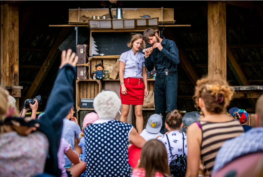Zdjęcie przedstawie fragment ze spektaklu. Na zdjęciu znajdują się dwie osoby na scenie, oraz tłum pod nią.