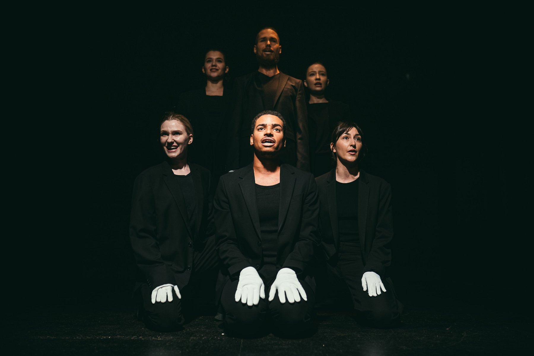 Trzy osoby siedzące obok siebie, w czarnych ubraniech i z białymi rękawiczkami na rękach. Za nimi ich odbicie. Czarne tło.