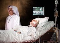 na łóżku szpitalnym leży męzczyzna podłączony do aparatury medycznej, obok stoi siostra zakonna