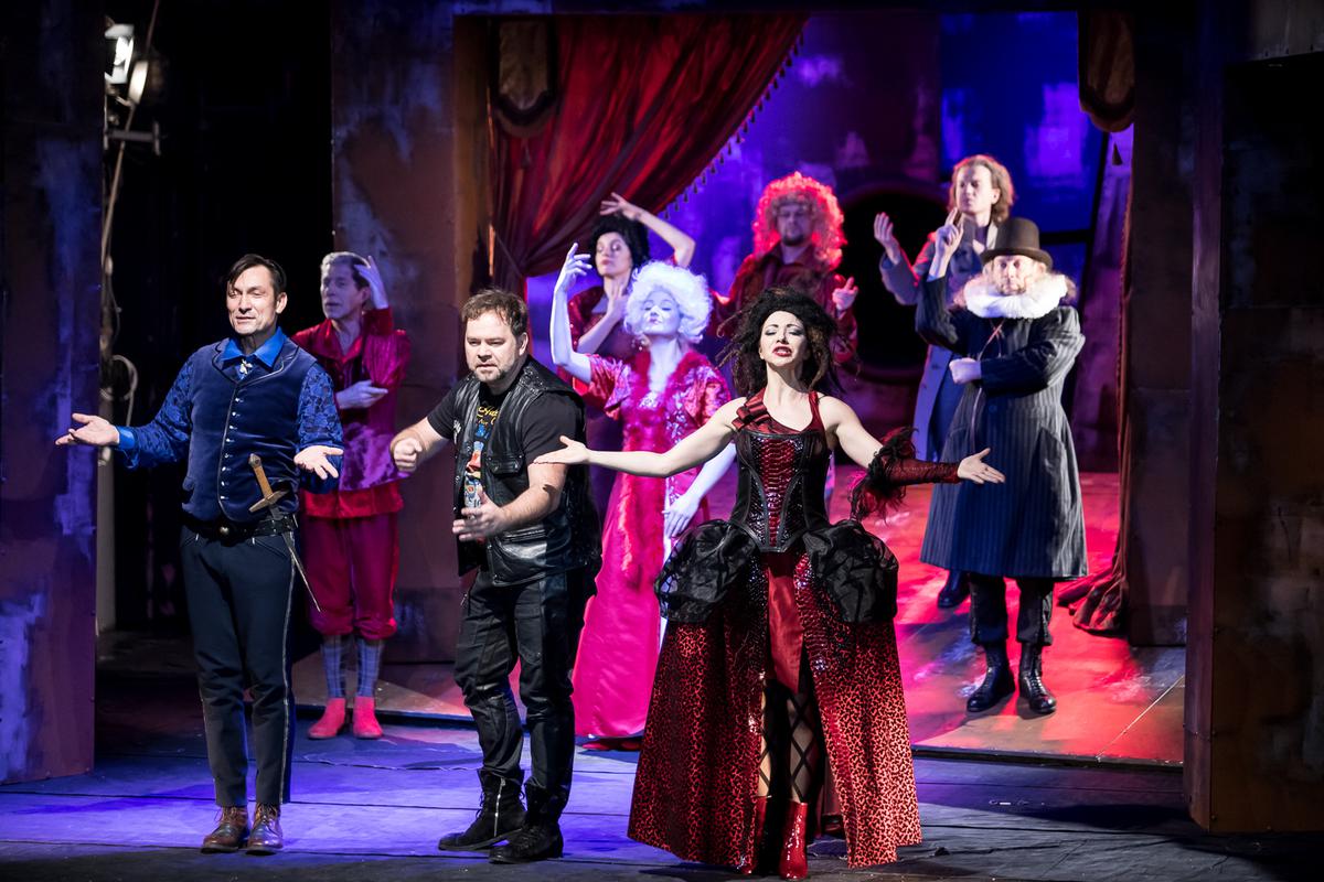 Zdjęcie ze spektaklu - na scenie aktorzy w różnorodnych strojach; na pierwszym planie aktorka w długiej balowej sukni, obok niej mężczyzna w skórzanych spodniach, skórzanej kamizelce i T-shircie.