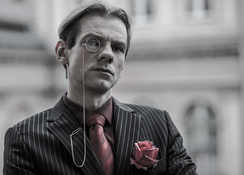 Czarno-białe zdjęcie mężczyzny w koszuli, krawacie i marynarce w paski, z monoklem przy oku. W kieszonce marynarki róża w różowo-szarym kolorze; w tym samym kolorze krawat i koszula mężczyzny.