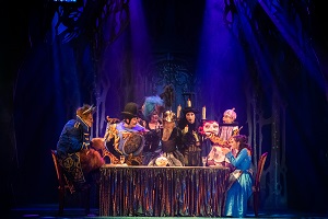 Zdjęcie ze spektaklu w granatowo-czarnych barwach; na pierwszym planie oświetleni aktorzy na scenie siedzący lub stojący wokół stołu.