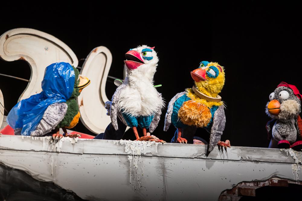 Na zdjęciu trzy kukuiełki kolorowych ptaków siedzacych na szarym murku.