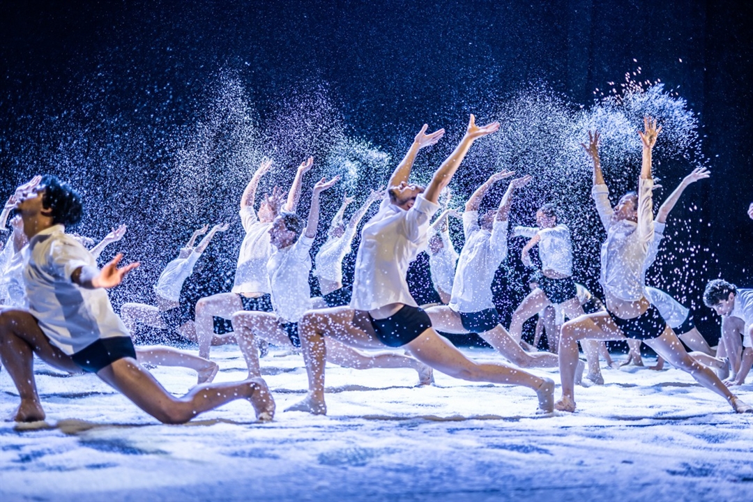 Tancerze na scenie w tanecznych pozach, wszyscy ubrani w białe koszule i czarne, krótkie spodenki. Wokół nich drobny pył przypominający śnieg.