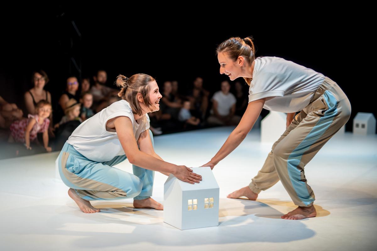 Dwie uśmiechnięte kobiety na scenie na przeciwko siebie, jedna stojąc pochyla się do przodu, druga kuca, obie dotykają białego, kartonowego domku. W tle publicznośc.