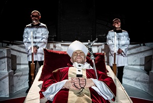 Mężczyzna ubrany w szaty papieskie leży na katafalku, pod głową ma purpurową poduszkę; za nim stoją dwaj mężczyźni w komżach.