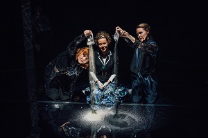 Trzy kobiety, dwie z nich wysypują piasek z woreczków na czarną powierzchnię. Czarne tło.