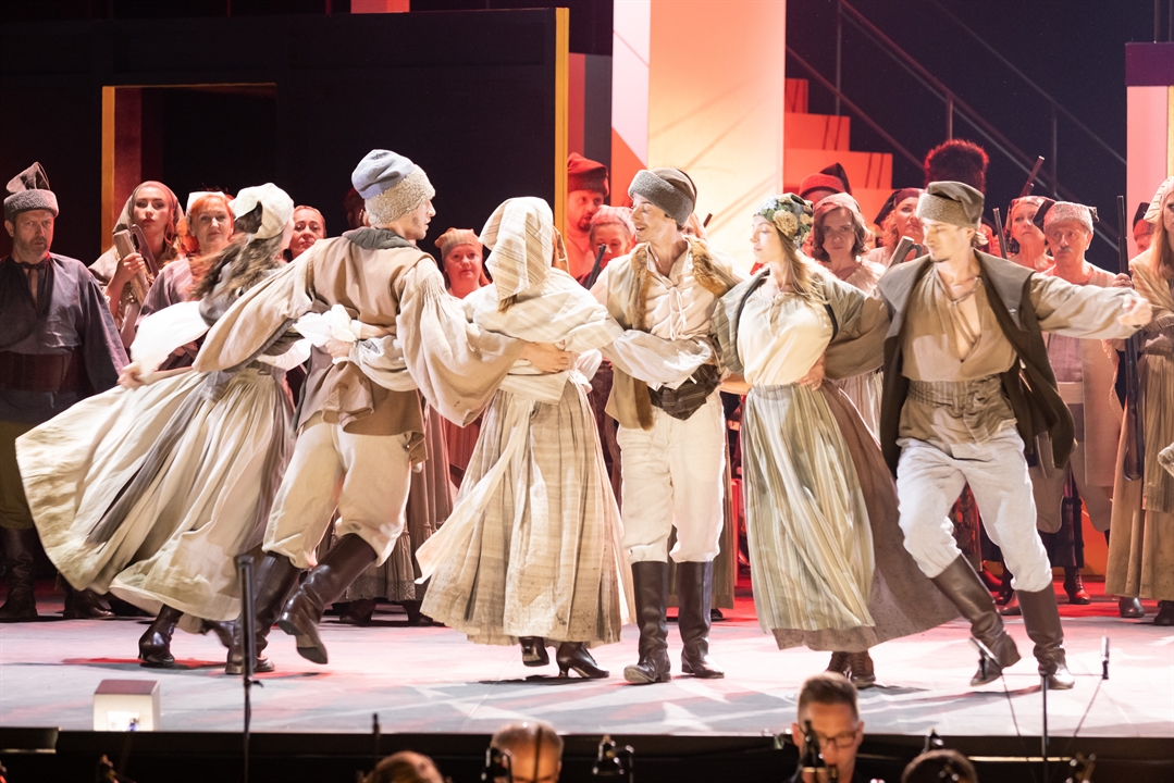 Grupa tancerzy w jasnych ludowych strojach tańczą trzymając się za ręce, w tle aktorzy, którzy patrzą na tańczących.