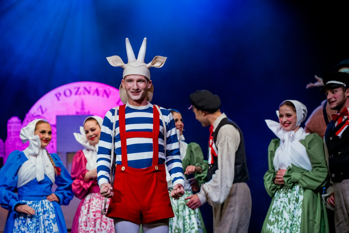 Aktor przebrany za Koziołka Matołka, w czerwonych spodniach na szelkach i biało-niebieskiej bluzce, za nim kilka osób w strojach ludowych.