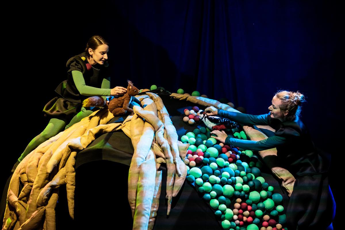 Na zdjęciu dwie kobiety, jedna trzyma pluszową wiewiórkę, druga - dzięcioła; obie aktorki stoją po dwóch stronach półkolistej konstrukcji złożonej z pasów materiału i kolorowych kulek.