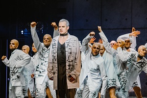 Zdjęcie z przedstawienia: grupa aktorów w białych strojach w różnych pozach na scenie. Na pierwszym planie wyprostowany mężczyzna.