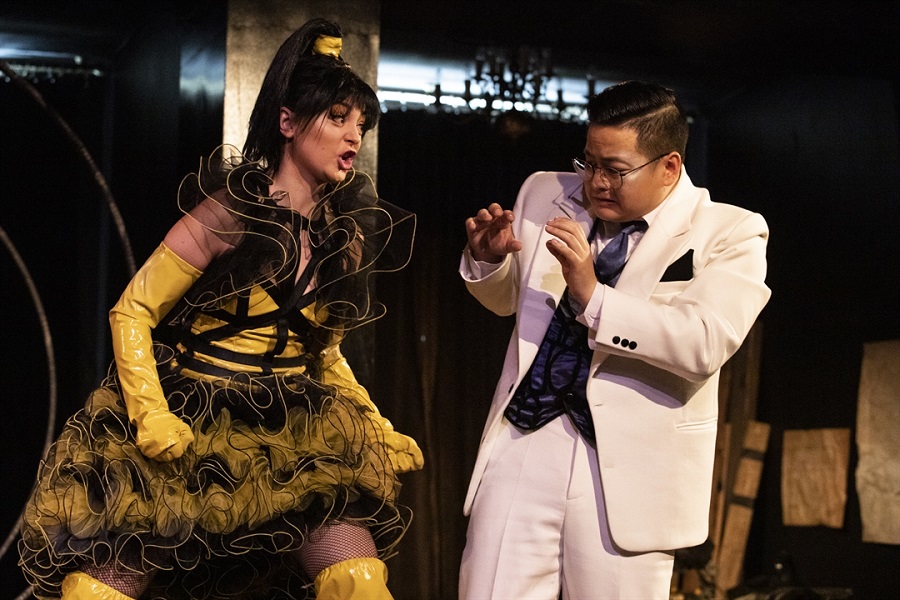 na zdjęciu mężczyzna ubrany w biały garnitur stoi unosząc ręce przed siebie przed krzyczącą na niego kobietą, ubraną w żółto-czarną sukienkę