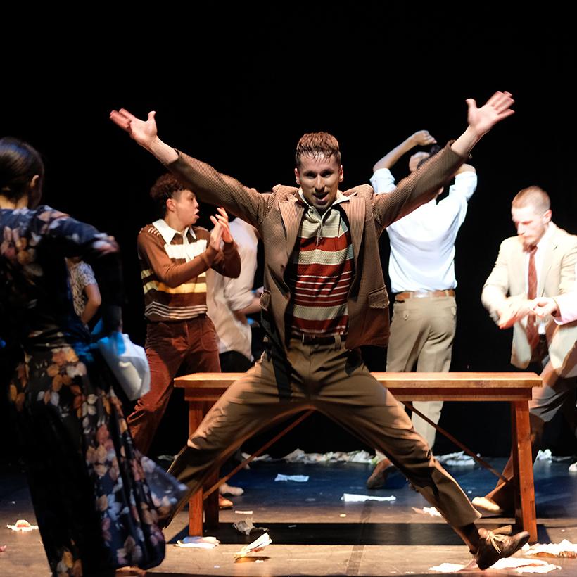 Mężczyzna z uniesionymi i wyprostowanymi rękoma, stojący w rozkroku, za nim drewniana ławka bez oparcia i kilku innych mężczyzn w różnych pozach.