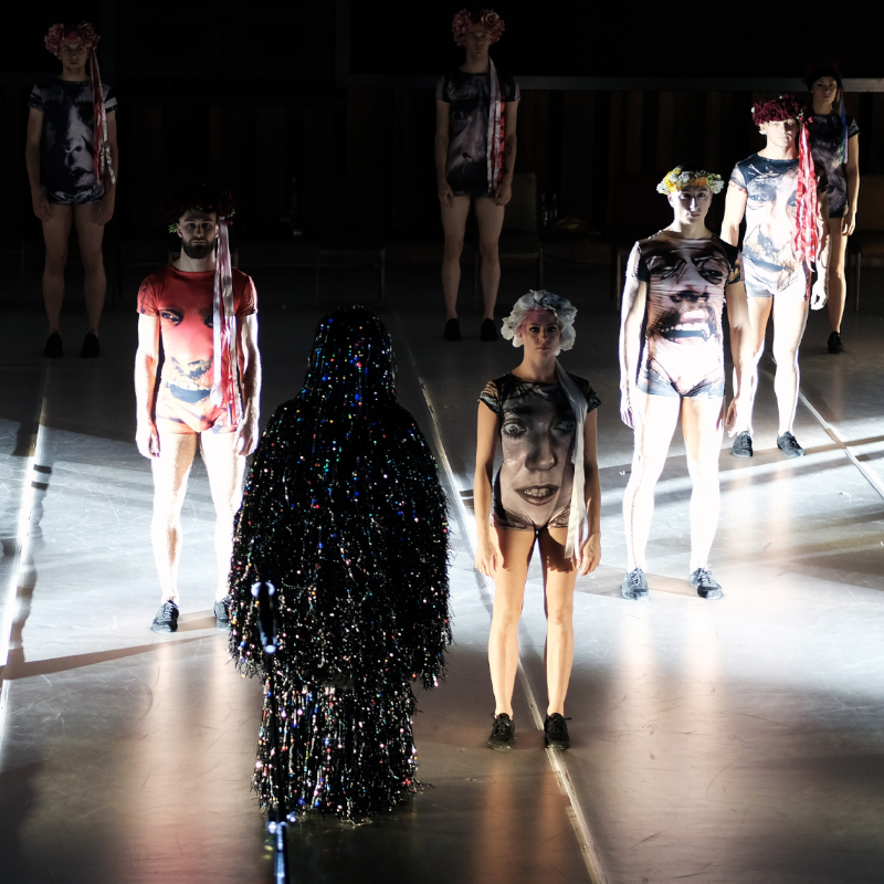 Kilka postaci stojących w pewnej odległości od siebie, w strojach z nadrukowanymi twarzami. Na pierwszym planie postać pokryta sznurami małych, świecących, kolorowych światełek.