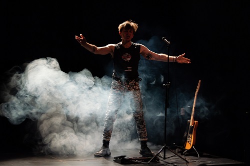 Mężczyzna stojący przy mikrofonie z rozpostartymi rękoma. Z boku stoi na stojaku gitara. Zdjęcie w półmroku, mężczyznę częściowo otacza biały dym.