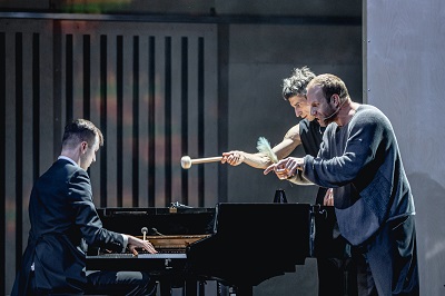 Na scenie trzech mężczyzn: jeden gra na fortepianie, dwaj pozostali stoją przy fortepianie zwróceni w kierunku pianisty. Jeden ze stojacych mężczyzn trzyma w dłoni pałeczke do gry na bębnach.