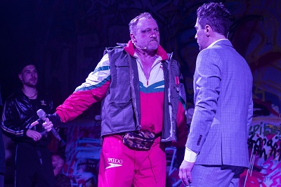 Dwóch mężczyzn stojących naprzeciwko siebie, jeden eleganco ubrany w garnitur, drugi w sportowym dresie i ortalionowej kamizelce.