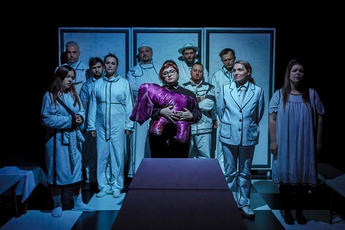 Na środku kobieta trzymająca fioletową przytulankę, za nią stoi dziesięć osób w białych ubraniach. Zdjęcie w niebiesko-białych kolorach.