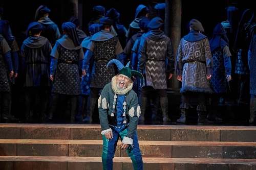 Błazen w zielono-niebieskim stroju i błazeńskiej czapce na scenie, za nim odwróceni tyłem do niego aktorzy, wszyscy w podobnych strojach i nakryciach głowy.