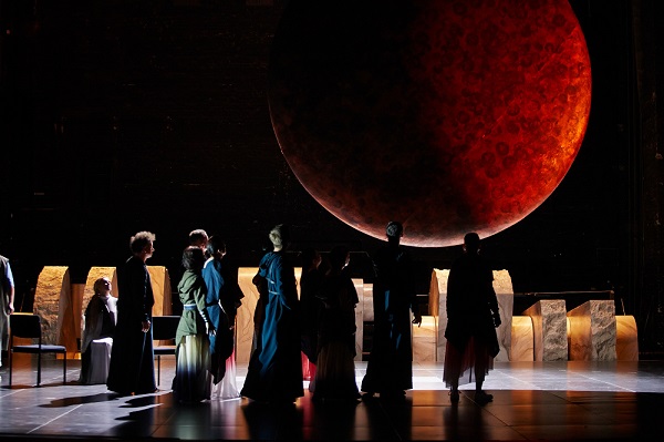 Ciemne zdjęcie sceny, nad którą unosi się duże czerwono-czarne koło przypominające księżyc. Na scenie aktorzy odwróceni tyłem do widzów i zwróceni w kierunku czerwonego koła. W tle elementy scenografii.
