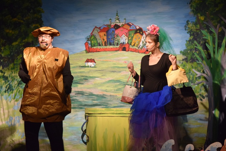 Mężczyzna ubrany w brązowy kostium, w niedużej odległości od niego kobieta trzymajaca kilka toreb, która patrzy na mężczyznę. W tle m.in. rysunek kilku domków otoczonych niewysokim murem.