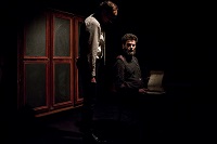 Ciemne zdjęcie: dwóch mężczyzn, jeden siedzi , drugi przy nim stoi. W tle drzwi szafy.