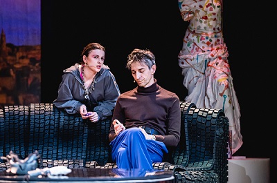 Mężczyzna siedzi na kanapie, za kanapą kobieta, która jest o nią oparta i skierowana w stronę mężczyzny. W tle fragment manekinu, na którym wisi kolorowa suknia.