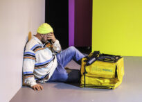 na zdjęciu mężczyzna siedzi pod ścianą, ręką zasłania twarz, przed nim na podłodze leży żółta torba termiczna do przewozu jedzenia