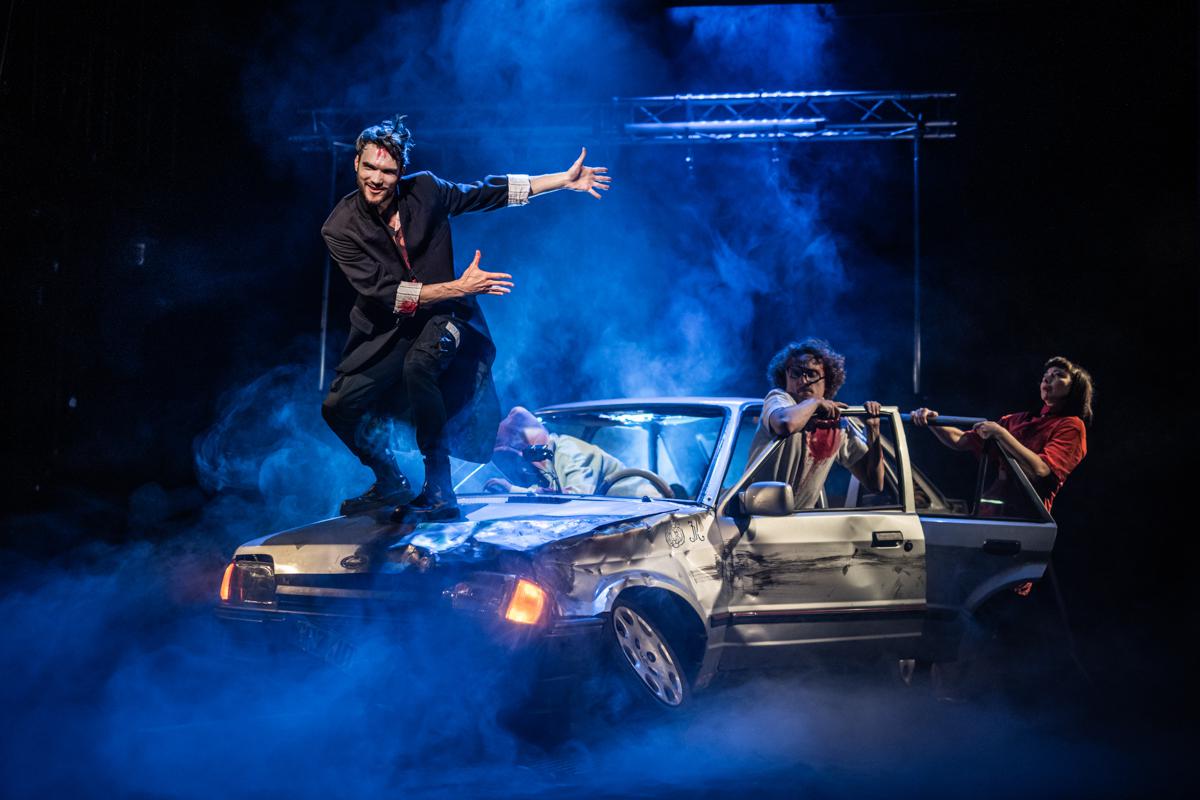 Zdjęcie przedstawiające trzy osoby wysiadające ze zniszczonego samochodu oraz jednego mężczyznę stojącego na masce samochodu.