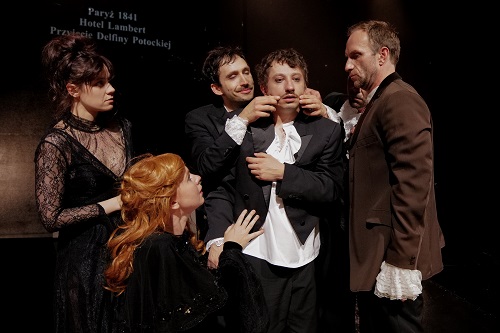 Dwóch mężczyzn soi blisko trzeciego mężczyzny, jeden z nich dotyka jego twarzy. Dwie kobiety przygladają się mężczyznom.