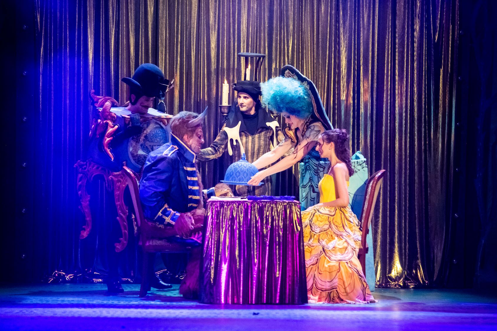 Przy stoliku siedzi kobieta w żółtej sukni, naprzeciw niej siedzi postawny mężczyzna w granatowym stroju, z rogami na głowie. Obok stołu stoją trzy postacie w strojach scenicznych, jedna z nich trzyma nad stolikiem półokrągłe naczynie.